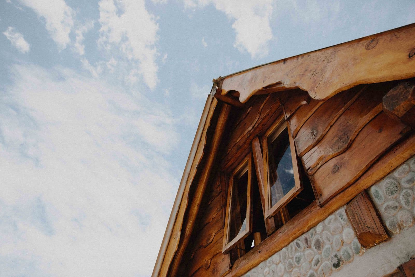 Close-up of a rustic wooden cabin with open windows against a blue sky with clouds.