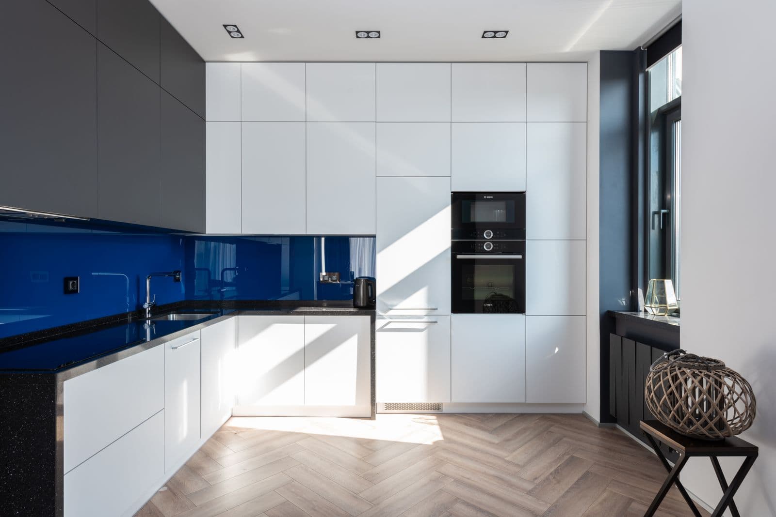 Interior of stylish kitchen with white and black cupboards with modern oven placed on parquet in light spacious apartment with decorations